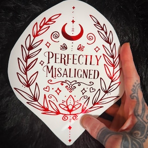May include: A white die-cut sticker featuring a metallic red design of a crescent moon, laurel branches, and decorative flourishes. The text "PERFECTLY MISALIGNED" is printed in the center in a bold, serif font, surrounded by small stars and hearts.