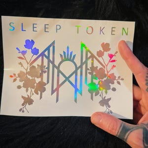 May include: A rectangular white sticker featuring the text SLEEP TOKEN in holographic lettering at the top. Below the text is a central geometric emblem flanked by detailed floral illustrations on both sides, all rendered in a reflective holographic finish.