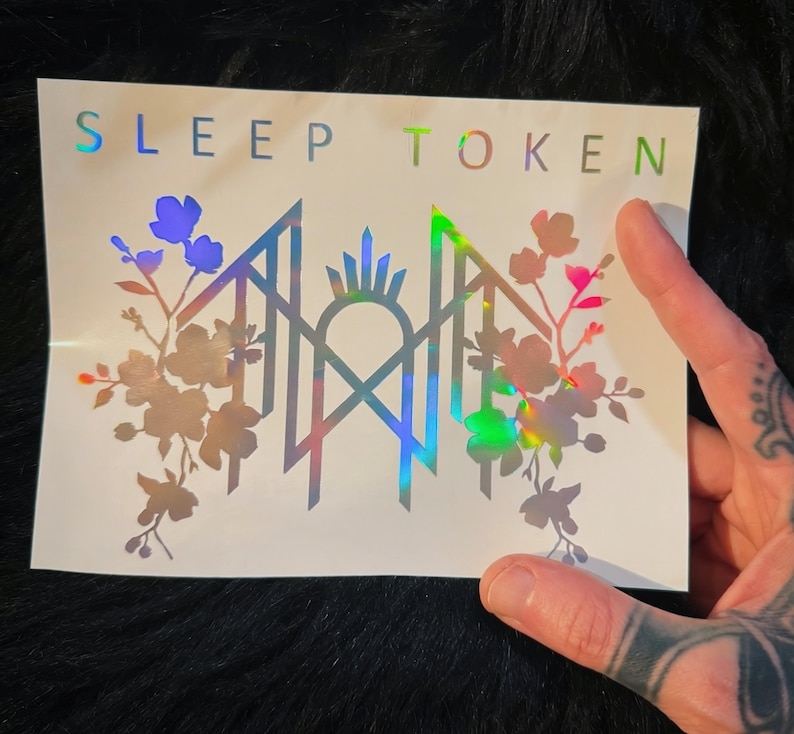May include: A rectangular white sticker featuring the text SLEEP TOKEN in holographic lettering at the top. Below the text is a central geometric emblem flanked by detailed floral illustrations on both sides, all rendered in a reflective holographic finish.