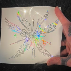 May include: A holographic vinyl decal featuring a central eye surrounded by four feathered wings, radiating starburst lines, and small star icons. The design includes a thin line extending to a small heart shape, all rendered in a reflective finish.