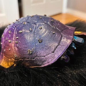 May include: A handcrafted, decorative turtle shell in shades of purple, blue, and gold. The shell is adorned with small, gold-colored embellishments and features a cluster of colorful crystals at the edge. The turtle shell is resting on a black, textured surface.
