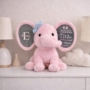May include: A plush pink elephant toy with a light blue bow on its head. The left ear features the letter E and the name Eleni. The right ear displays the date 08.03.2025, the weight 7 pounds 12 oz, and the time 2:38am with baby footprint icons.