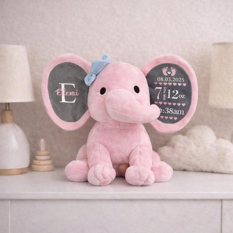 May include: A plush pink elephant toy with a light blue bow on its head. The left ear features the letter E and the name Eleni. The right ear displays the date 08.03.2025, the weight 7 pounds 12 oz, and the time 2:38am with baby footprint icons.