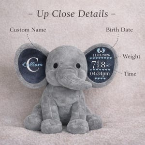 May include: A gray plush elephant toy featuring dark blue inner ears. The left ear displays the name Callum in white script. The right ear shows birth details including the date 11.03.2026, weight 7 pounds 8 ounces, and time 04:34pm.
