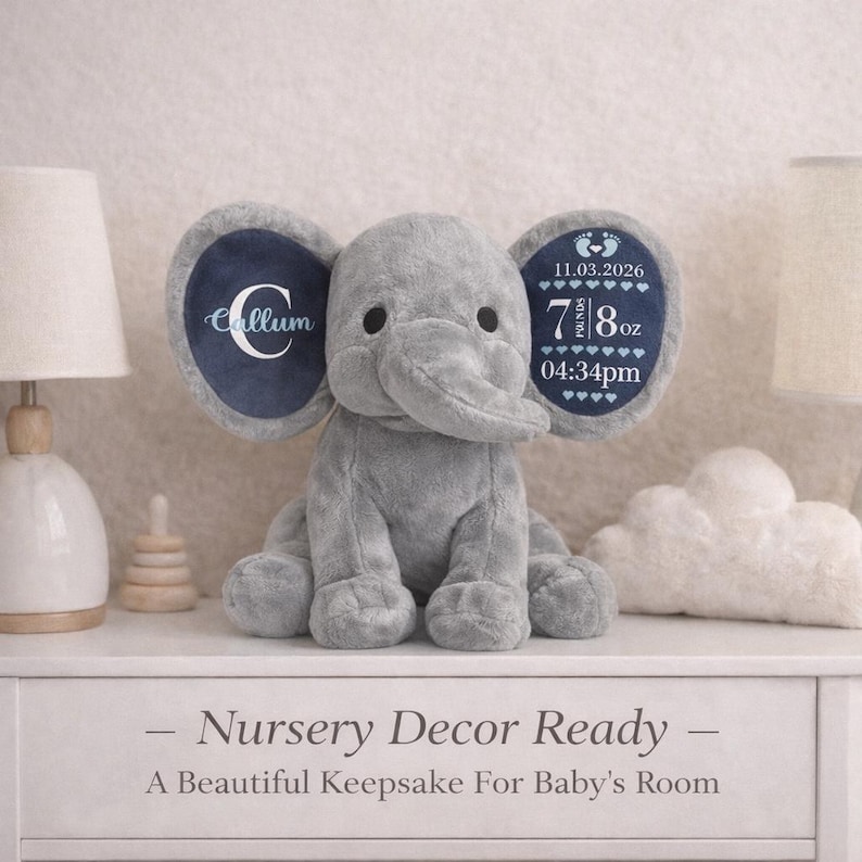 May include: A grey plush elephant toy with large ears. The left ear features a white letter C and the name Callum. The right ear displays the date 11.03.2026, the weight 7 lbs 8 oz, and the time 04:34pm with small heart icons and baby footprints.