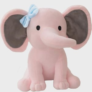 May include: A plush elephant toy in a soft pink color with large ears featuring dark gray inner panels. The elephant has small black eyes and a light blue fabric bow attached to the top of its left ear. It is depicted in a seated position.