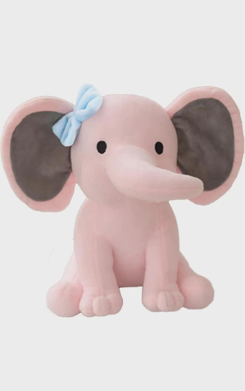 May include: A plush elephant toy in a soft pink color with large ears featuring dark gray inner panels. The elephant has small black eyes and a light blue fabric bow attached to the top of its left ear. It is depicted in a seated position.