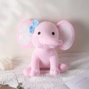 May include: A soft plush elephant toy in a light pink color with large ears featuring white inner sections. The elephant has two small black eyes and a light blue fabric bow attached to the top of its left ear. The toy is in a seated position.