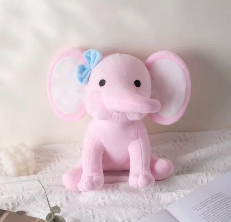 May include: A soft plush elephant toy in a light pink color with large ears featuring white inner sections. The elephant has two small black eyes and a light blue fabric bow attached to the top of its left ear. The toy is in a seated position.