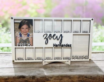 May include: White wooden photo frame with a stand, designed to hold photos from each year of school. The frame has slots for photos from pre-K through senior year. The frame has the name "Zoey Hernandez" written on it. A photo of a young girl is in the pre-K slot.