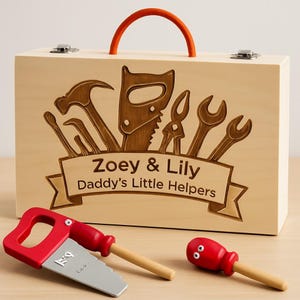 May include: A wooden toy toolbox with an orange handle and metal clasps. The front of the box features engraved tools and the text "Zoey & Lily Daddy's Little Helpers." A red toy saw and screwdriver are in front of the box.