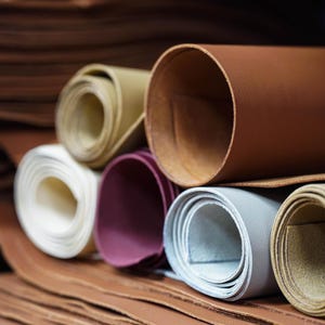 May include: A selection of rolled leather hides in a variety of colours, including brown, white, purple, and grey. The image suggests the leather is of good quality, suitable for crafting.