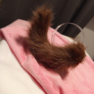May include: A brown, furry tail with a clear, circular attachment, resting on a pink, plush fabric. The tail is curved and appears to be made of soft, textured material. The background is a soft, neutral color.