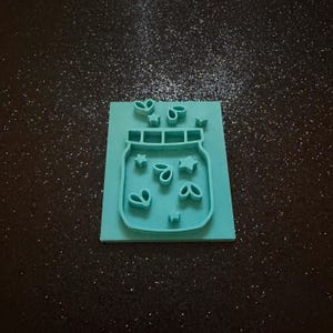 May include: A light teal rectangular cookie cutter featuring a mason jar design. The interior of the jar is embossed with small star and leaf shapes, while additional star and leaf shapes are positioned above the jar outline on the base.