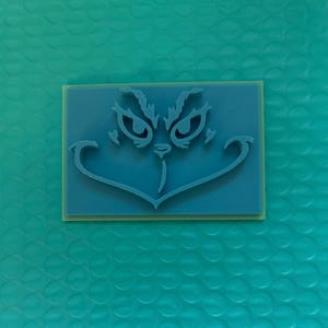 May include: A blue rectangular stamp featuring the Grinch's face. The design includes the Grinch's eyes, eyebrows, nose, and mouth. The stamp is set on a light yellow base, with a teal background.