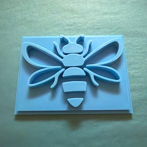 May include: A light blue silicone mold featuring a raised bee design. The mold has a rectangular base with a centered, stylized bee silhouette that includes detailed wings, a segmented body, and antennae, all in a solid light blue color.