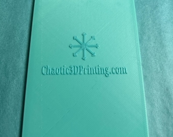 May include: A teal-colored 3D-printed scraper with a snowflake design and the text "Chaotic3DPrinting.com". The scraper has a textured surface and a unique shape with a wavy top and a scalloped bottom edge.