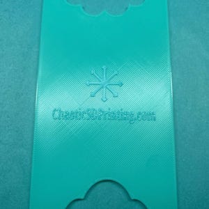 May include: A teal-colored, rectangular 3D-printed object with a decorative top and bottom edge. The object has a textured surface and features a snowflake design and the text "Chaotic3DPrinting.com".