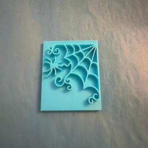 May include: A rectangular light blue silicone mold featuring a raised design of a spider web in the upper right corner and a small spider with curved legs on the left side. The mold has a smooth flat base and intricate cutouts for the web pattern.