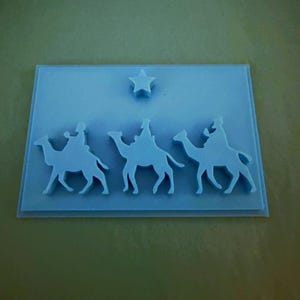 May include: A light blue rectangular silicone mold featuring a raised design of three figures riding camels in a row, with a single five-pointed star positioned above the center figure. The mold has a smooth, flat base and defined edges.