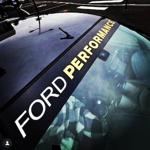 May include: Black car windscreen with a banner displaying "FORD PERFORMANCE" in white and gold lettering. The car's interior is visible, including a plaid blanket on the seat.