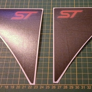 Pode incluir: Dois decalques ST pretos e cinzentos com uma borda branca. O logotipo ST é vermelho. Os decalques são triangulares e estão sobre uma base de corte verde.