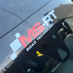 May include: A black vehicle windshield featuring a white and red decal that reads MS-RT ROAD TECHNOLOGY. The design includes a white checkered pattern element to the left of the red letters MS, followed by white letters RT and text below.