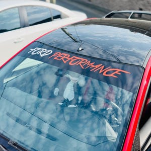 Peut inclure: Une voiture rouge et noire avec une bannière de pare-brise indiquant "FORD PERFORMANCE" en lettres blanches et rouges. L'intérieur de la voiture est visible à travers le pare-brise, et une autre voiture blanche est garée à proximité.