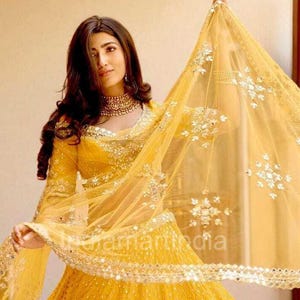May include: A bright yellow lehenga, a traditional Indian outfit, adorned with detailed mirror work and embellishments. The ensemble features a long-sleeved top and a matching dupatta. The dupatta is draped, highlighting the delicate embroidery. The text "theamaraindia" is visible.