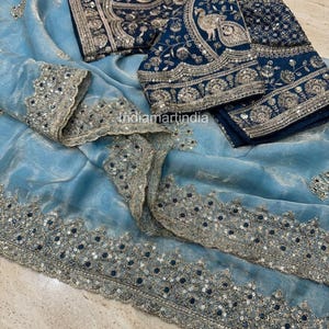 May include: A light blue saree with intricate silver embroidery and embellishments. The saree is paired with a matching navy blue blouse, also featuring detailed silver embroidery and a peacock design. The fabric appears soft and flowing.