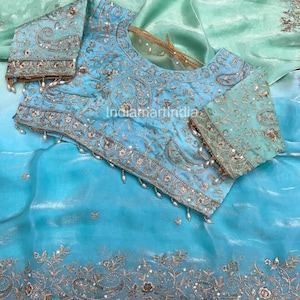 May include: A light blue embroidered blouse and matching sheer fabric. The blouse features intricate silver and gold embroidery with pearl accents. The sheer fabric has a floral embroidered border. The overall design is elegant and detailed.