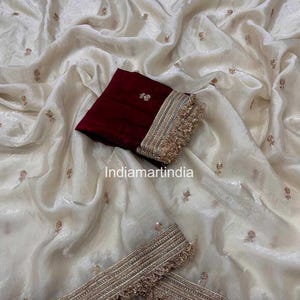 Puede incluir: Sari de color marfil con delicados bordados florales y ribetes dorados. Se incluye una pieza de blusa de terciopelo rojo oscuro con bordados a juego y flecos dorados. El sari tiene un aspecto brillante y texturizado.