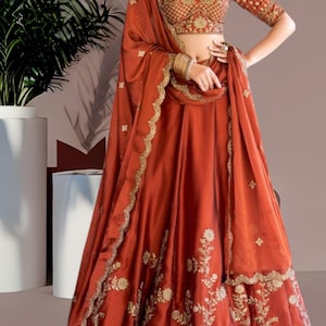 Könnte beinhalten: Ein rostfarbener Seidenlehenga mit goldener Stickerei. Der Rock hat eine ausgestellte Silhouette und ist mit Blumenmustern verziert. Die Dupatta ist ebenfalls rostfarben und hat einen goldenen Rand.