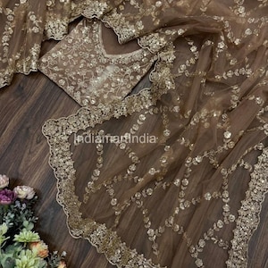 Puede incluir: Conjunto de tela marrón transparente con intrincados bordados dorados y lentejuelas. El conjunto incluye una blusa y un chal drapeado, ambos con motivos florales y un borde festoneado. La tela está dispuesta sobre una superficie de madera oscura.