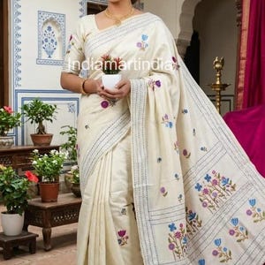 May include: Ivory-colored sari with floral embroidery in blue, purple, and pink. The sari features a detailed border and is worn with a matching blouse. The text "indiamartindia" is visible on the sari.