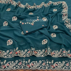 Puede incluir: Sari de color verde azulado con bordados intrincados. El sari presenta patrones florales y de cachemira detallados en rojo, dorado y blanco. Incluye una pieza de blusa a juego con bordados similares. El texto "IndiamartIndia" es visible en la tela.