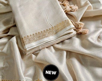 Puede incluir: Tela color crema con un patrón de cuadrícula y bordes decorativos dorados. La tela está doblada, con borlas visibles. Un círculo negro con la palabra "NEW" está en la imagen.