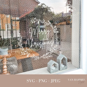 May include: White text on a window that reads "Merry Christmas" in a wreath of white leaves. The window is behind a wooden table and two small grey houses.