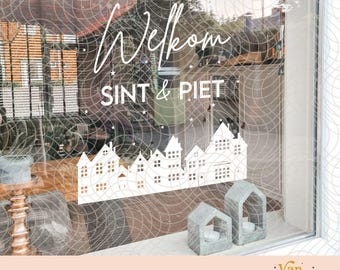 May include: A white vinyl window decal featuring a row of houses and the text Welkom SINT & PIET. The design includes small stars and a cityscape silhouette. Two small house-shaped candle holders are placed on the window sill below the decal.