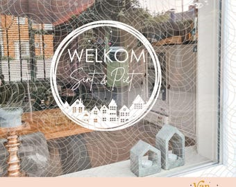 May include: A white circular window decal featuring the text WELKOM Sint & Piet above a silhouette of a row of houses. The design is applied to a glass surface, with two small house-shaped candle holders visible on the ledge below the decal.