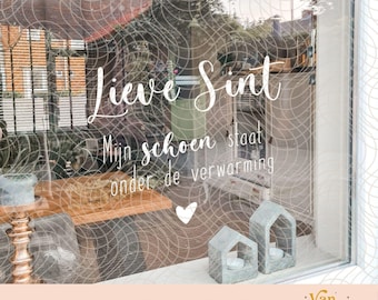 May include: A white vinyl window decal featuring the Dutch text Lieve Sint Mijn schoen staat onder de verwarming with a small heart icon. The design is displayed on a glass surface with a background view of a house and two small house-shaped candle holders.