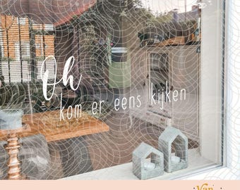 May include: A window decal featuring the white cursive text "Oh kom er eens kijken" applied to a glass surface. The decal is displayed on a window looking out onto a patio with a wooden table, two small house-shaped concrete candle holders.
