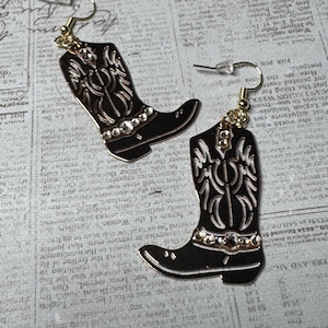 May include: A pair of dangling earrings shaped like cowboy boots. The boots are black with gold-toned outlines and decorative cutouts. Each boot features a row of small, clear rhinestones across the ankle area and hangs from a gold-toned hook.