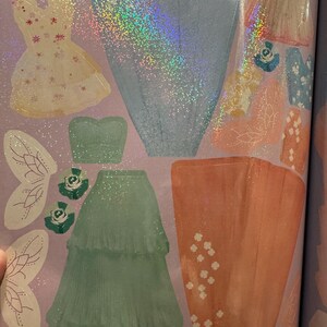 May include: A selection of dress-up stickers, showcasing diverse dress designs. These include a pale yellow dress with floral details, a shimmering blue dress, and a tiered, mint green skirt. Fairy wings and floral decorations are also present.