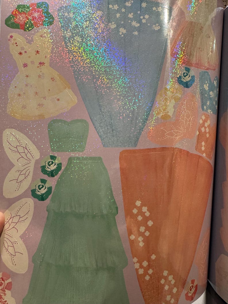 May include: A selection of dress-up stickers, showcasing diverse dress designs. These include a pale yellow dress with floral details, a shimmering blue dress, and a tiered, mint green skirt. Fairy wings and floral decorations are also present.