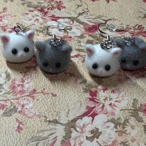 May include: Four cat-shaped earrings. Two pairs of earrings are visible, each featuring a white or gray cat face with pink inner ears and black eyes. The earrings are on silver-colored hooks. The background is a floral pattern.