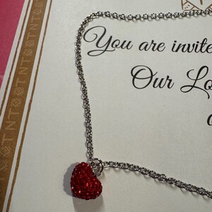 May include: A silver chain necklace featuring a small, red heart pendant. The heart is adorned with tiny, sparkling red stones. The background includes the words "You are invited".