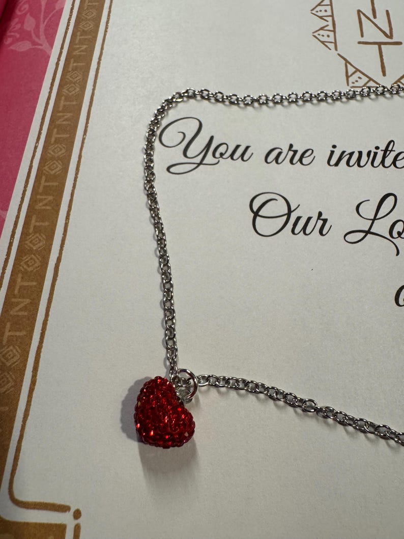 May include: A silver chain necklace featuring a small, red heart pendant. The heart is adorned with tiny, sparkling red stones. The background includes the words "You are invited".