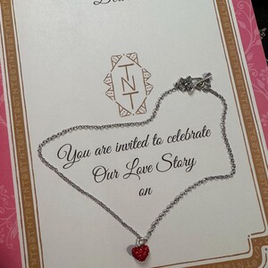 May include: A silver necklace with a small, red heart pendant is displayed on a greeting card. The card features decorative borders and the text "Dear" and "You are invited to celebrate Our Love Story on."