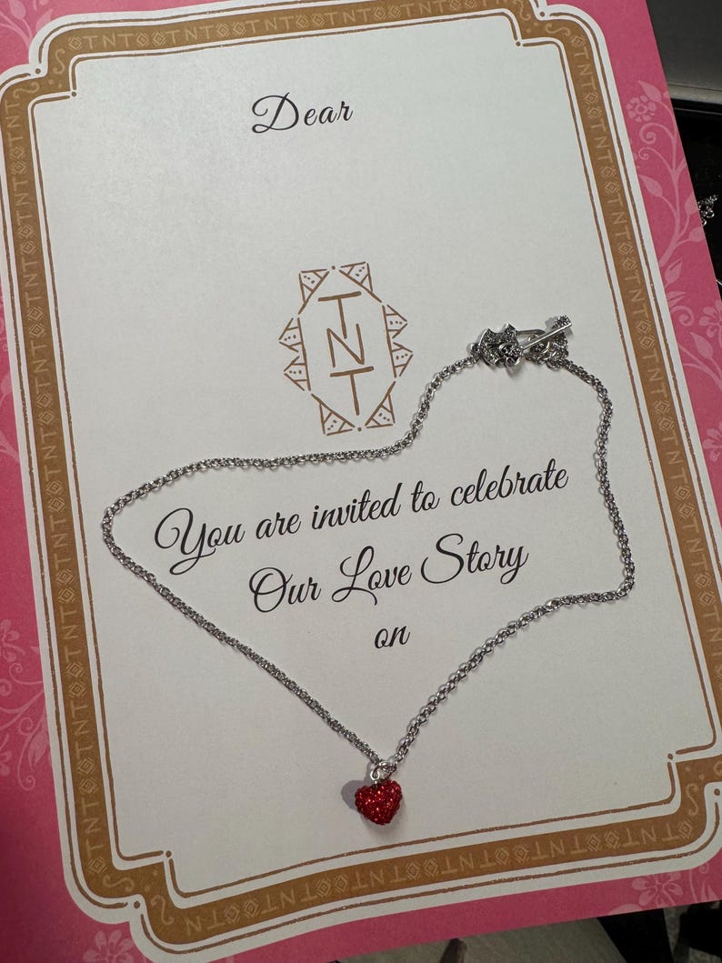 May include: A silver necklace with a small, red heart pendant is displayed on a greeting card. The card features decorative borders and the text "Dear" and "You are invited to celebrate Our Love Story on."